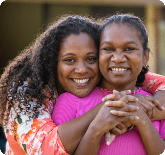 Two young aboriginal female students outdoors with their arms around each other smiling at the camera.