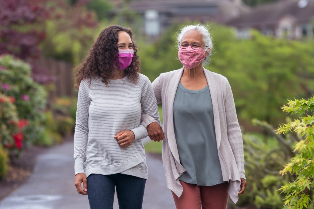 Two mixed race women wearing protective face masks and are walking with their arms linked. They are enjoying the outdoors and getting some exercise during COVID-19.