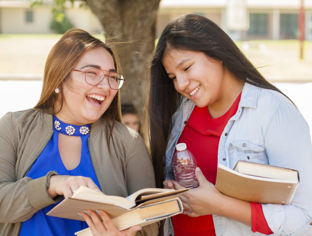 Multi-ethnic group of high school girls holding books talking together on campus outdoors.